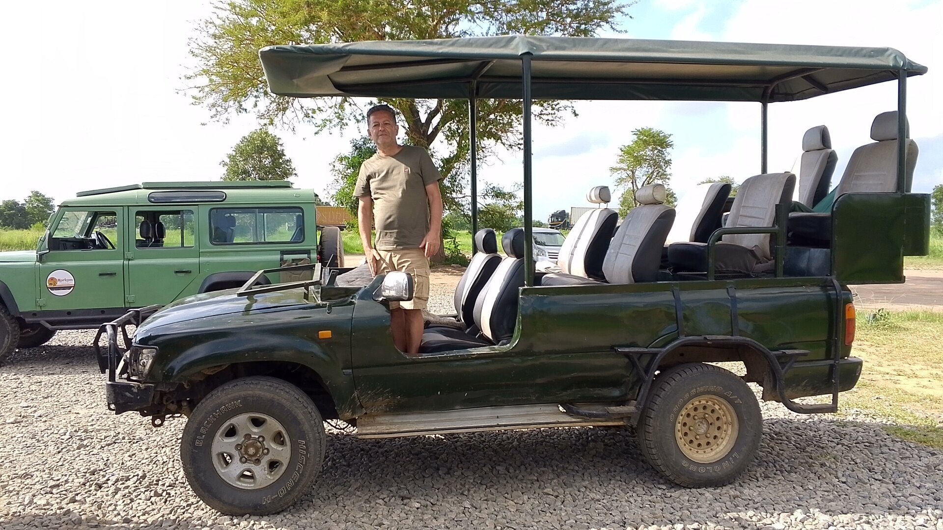 A guide standing beside an open-sided green safari truck on gravel in Mikumi